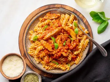 Pyszny makaron z kurczakiem w sosie pomidorowym, udekorowany bazylią. Obok miseczki z parmezanem i ziołami.