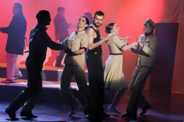 Aktorzy w strojach z epoki tańczą na scenie z czerwonym tłem.