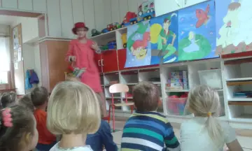 Nauczycielka w czerwonym kapeluszu i stroju opowiada bajkę, trzymając koszyczek Czerwonego Kapturka. Dzieci słuchają z uwagą.