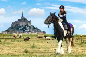 Une jeune fille rit à cheval devant le Mont-Saint-Michel, avec des moutons dans un pré.