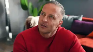 Justin z "Barw Szczęścia" w czerwonym swetrze, zamyślony, w tle kanapa z kolorowymi poduszkami.