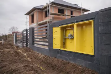 Żółta skrzynka z instalacją gazową w domu w budowie, obok ogrodzenia i placu budowy.