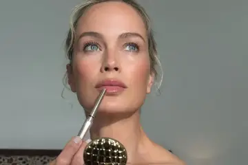 Kobieta z błękitnymi oczami aplikuje kredkę do ust, tworząc makijaż krok po kroku. Złote lusterko czeka na swoją kolej.