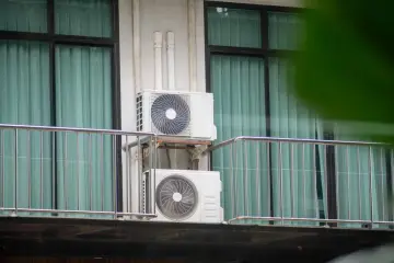 Dwa białe klimatyzatory zamontowane na ścianie budynku, na tle okien z zielonymi zasłonami.