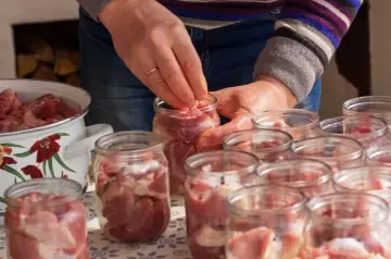 Ręce wkładają kawałki mięsa do słoików, przygotowując domowy gulasz angielski. Obok stoi garnek z pokrojonym mięsem.