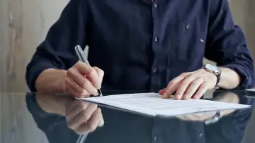 Mężczyzna w granatowej koszuli podpisuje aneks do umowy o pracę. Na ręce ma zegarek.