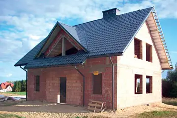 Budowa domu z cegły, widoczny dach betonowy skośny ocieplenie. Widać otwory okienne i drzwiowe, komin.