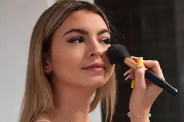 Młoda kobieta z długimi blond włosami nakłada puder pędzlem. Jej makijaż jest subtelny, podkreślający naturalne piękno, w stylu clean girl.