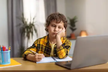 Chłopiec z czwartoklasistą znużony pisze wypracowanie. Na biurku laptop, zeszyt i przybory.