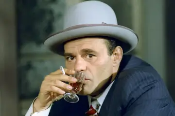 Nikodem Dyzma w kapeluszu, popijający drinka i palący papierosa. W jego oczach widać pewność siebie i przebiegłość.