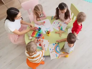 Dzieci rysują przy stole z nauczycielką. Zastanawiasz się, ile wynosi subwencja na dziecko w przedszkolu?