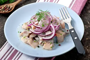 Sałatka śledziowa z cebulą i koperkiem na jasnoniebieskim talerzu, obok widelec.