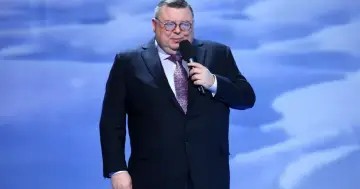 Mężczyzna w garniturze i okularach przemawia na scenie podczas turnieju piosenki.