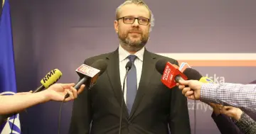 Marcin Wojciechowski w okularach, w garniturze, otoczony przez mikrofony mediów, udziela wywiadu.