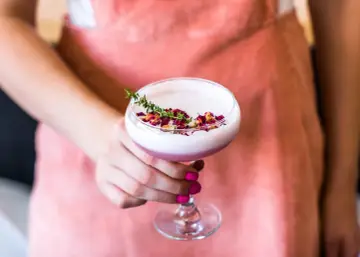 Różowy koktajl Pink Lady ozdobiony płatkami róż i gałązką ziół, trzymany w dłoni.