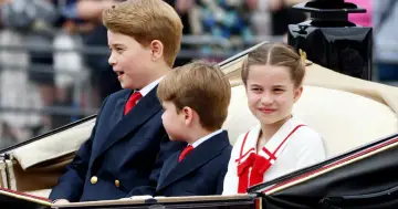 Dzieci Williama i Kate w karocy: książę George, książę Louis i księżniczka Charlotte, uśmiechnięta Charlotte w białej sukience z czerwoną kokardą.