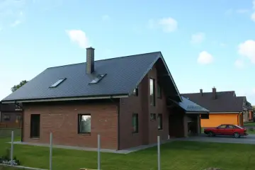Nowy dom z cegły, z ciemnym dachem i kominem o imponującej wysokości. W tle inne domy i czerwony samochód.