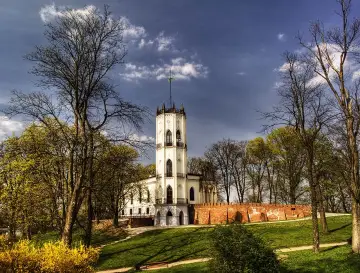 Biały zamek z wieżą na wzgórzu, otoczony drzewami i ceglanym murem. Wiosenna zieleń i żółte krzewy dodają uroku tej opinogórze.