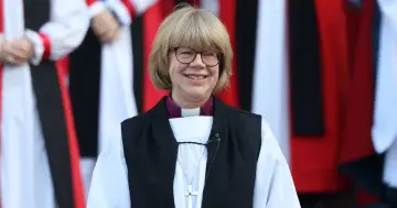 Uśmiechnięta kobieta w stroju duchownym, z krzyżem na szyi, przypomina arcybiskupa Canterbury.