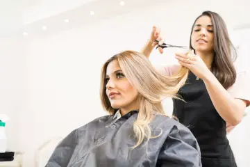 Fryzjerka przycina blond włosy klientki, tworząc nową, modną fryzurę. Efekt końcowy to ładnie obcięte włosy.