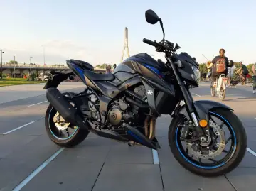 Czarny motocykl Suzuki GSX 750 na tle mostu. W tle ludzie spacerują i jeżdżą na rowerach.