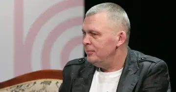 Adam z zespołu Raz Dwa Trzy w szarej marynarce, siedzi na kanapie.