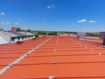 Pomarańczowy, lekki dach płaski z metalowymi elementami, rozciągający się pod błękitnym niebem z chmurami.