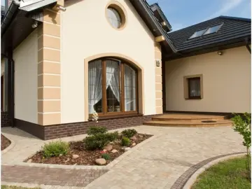 Elewacja domu kostka w jasnym kolorze, z ozdobnymi elementami i brązową cegłą u podstawy.