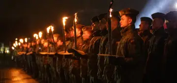 Żołnierze w czapkach typu capstrzyk niosą pochodnie podczas nocnej uroczystości.