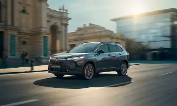 Ciemnoszary Toyota Corolla Cross sunie ulicą miasta, mijając zabytkowe budynki i nowoczesne szklane fasady.