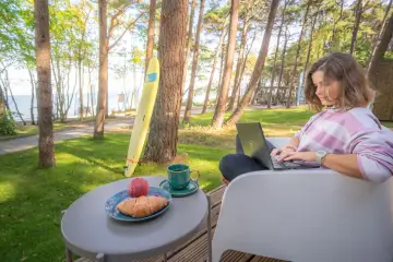 Kobieta pracuje na laptopie przy stoliku z kawą i croissantem. W tle widać morze i domki z wyjściem na plażę.