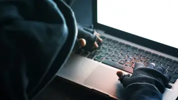 Osoba w kapturze i rękawiczkach pisze na laptopie. To może być początek kradzieży tożsamości.