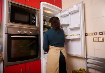 Kobieta w fartuchu zagląda do lodówki. Obok stoi piekarnik, a nad nim wbudowana mikrofalówka. Czy mikrofalówka może stać na lodówce? To pytanie, na które odpowiedź znajdziesz w instrukcji urządzenia.