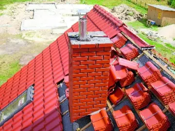 Trwa montaż dachówki, a komin czeka na obróbki komina blachą. Widać stosy dachówek i fundamenty nowego domu.