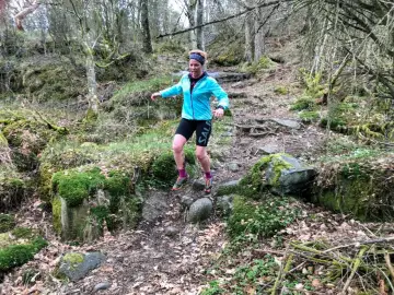 Kvinna springer i skogen, fokuserad på att springa marathon. Mossbeklädda stenar och träd skapar en utmanande terräng.