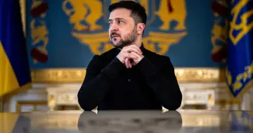 Prezydent Ukrainy Wołodymyr Zełenski w czarnej koszuli, z dłońmi złożonymi na stole, na tle flagi Ukrainy i herbu.