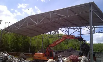 Solidna wiata stalowa rolnicza chroni plac budowy przed deszczem. Koparka z chwytakiem pracuje przy składowisku odpadów.