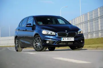 Ciemnoniebieski BMW van na tle jasnego nieba i barier dźwiękochłonnych.