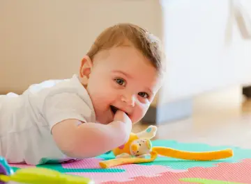 Uśmiechnięte 11 miesięczne dziecko leży na macie, gryząc paluszek. Obok pluszowa zabawka.