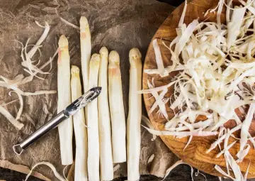 Frischer weißer Spargel wird geschält, die Spargelschalen liegen daneben. Ein Sparschäler liegt auf den Spargelstangen.