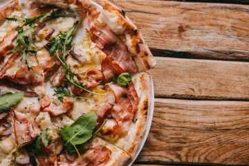 Pyszna pizza carbonara z chrupiącym boczkiem, rukolą i cebulą na drewnianym stole.