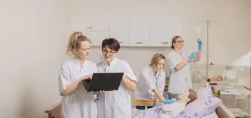 Pielęgniarki omawiają dane na laptopie, podczas gdy koleżanka ćwiczy zakładanie kroplówki. Zastanawiasz się, ile zarabia pielęgniarka po studiach magisterskich?