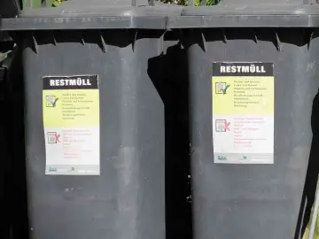 Dwa szare kosze na śmieci z etykietami "REST MÜLL". Etykiety zawierają informacje o tym, co można, a czego nie można wyrzucać. Wymiary kosza na śmieci są standardowe.
