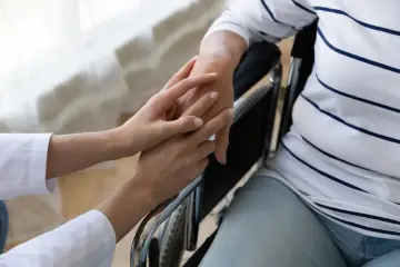 Dwie dłonie, jedna w białym rękawie, delikatnie dotyka drugiej, siedzącej na wózku. To obraz troski i wsparcia, być może świadczenie uzupełniające.