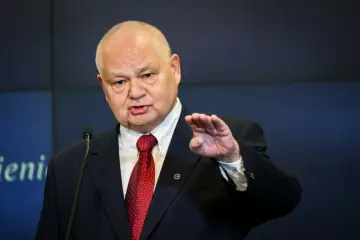 Prezes NBP w garniturze i czerwonym krawacie przemawia przy mikrofonie, gestykulując prawą ręką.