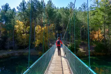 Senderista cruza puente colgante sobre el río, camino al Salto de Poveda y la Laguna de Taravilla. Bosque otoñal.