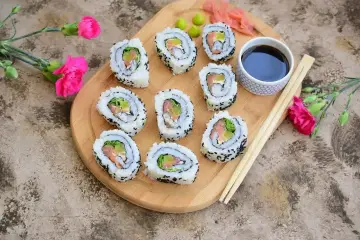 Pyszne california sushi z łososiem i awokado, podane z sosem sojowym i wasabi na drewnianej desce, ozdobione różowymi kwiatami.