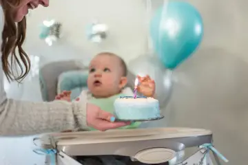 Ciocia podaje tort na roczek. Dziecko wpatrzone w świeczkę, balony i ozdoby tworzą radosną atmosferę.