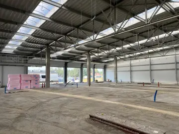 Wnętrze w budowie hali magazynowej. Betonowe słupy, stalowa konstrukcja dachu z świetlikami, ściany z płyt warstwowych. Na podłodze piasek i materiały budowlane.