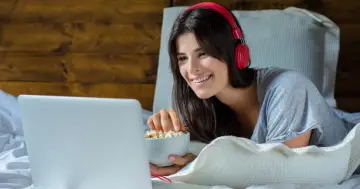 Uśmiechnięta dziewczyna w słuchawkach ogląda coś na laptopie, jedząc popcorn. To jej ulubiony sposób na relaks z multimedialnym odtwarzaczem.